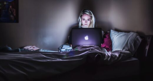 Woman sitting in the dark in bed, working on a laptop with headphones on