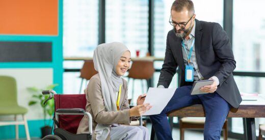 Asian Muslim businesswoman sitting on wheelchair presenting business graph on paper to manager. Smiling group of diverse corporate colleagues in the modern office. Diversity or multicultural in office