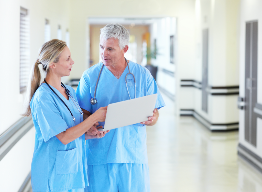 Two doctors in scrubs standing together and having a discussion over a laptop in a hospital corridor