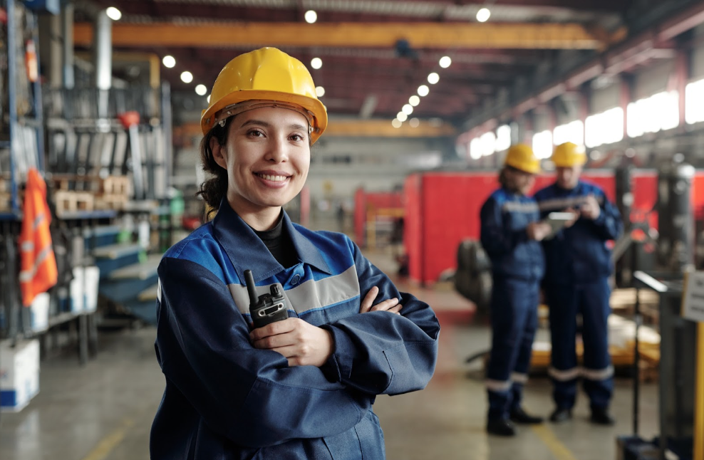 Young smiling female worker of modern industrial plant or factory in workwear and protective helmet standing in large workshop