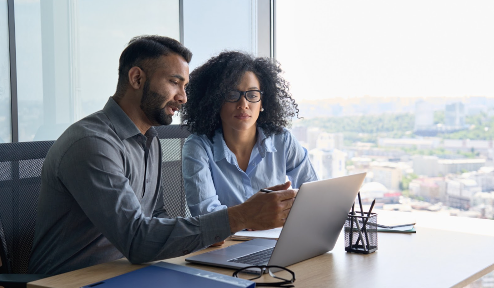 Indian male ceo executive manager mentor giving consultation on financial operations to female African American colleague intern using laptop sitting in modern office near panoramic window.