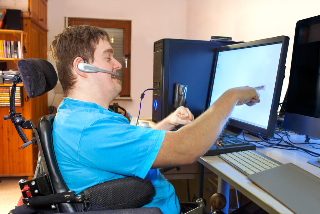 Young man with infantile cerebral palsy caused by a complicated birth sitting in a multifunctional wheelchair, using a computer with a wireless headset, reaching out to touch the touch screen.