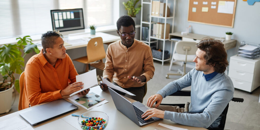 Portrait of diverse business team discussing project at meeting table focus on young man using wheelchair in foreground, inclusivity at workplace