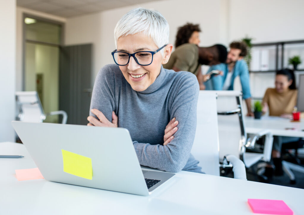 Woman looking at her work laptop while her colleagues discuss, joke and laugh.