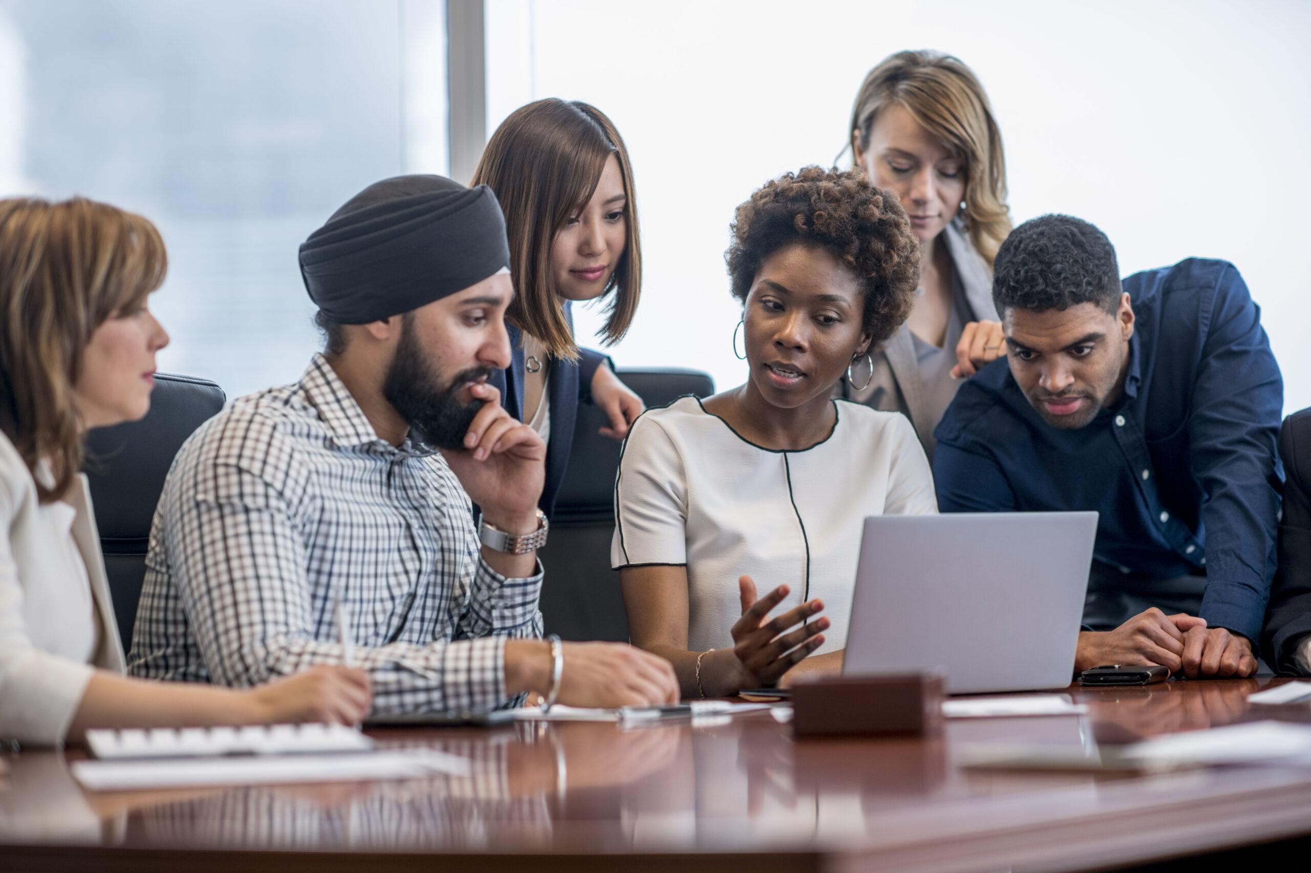 A diverse group of business people gather around a laptop in a modern office and discuss what they see.