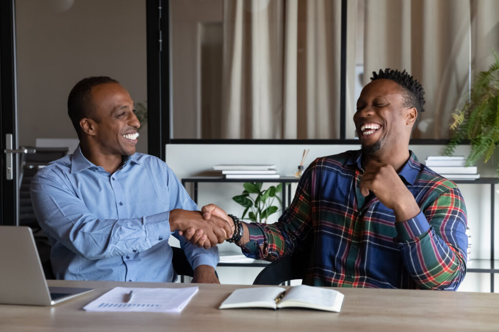 Overjoyed African American business partners shaking hands at meeting greeting or closing deal sitting at table in office, happy hr manager congratulating successful candidate after job interview