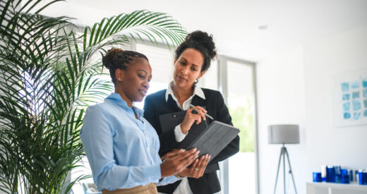African American businesswoman and a Hispanic female colleague sharing online documents with their coworkers. They are standing in a modern office space.