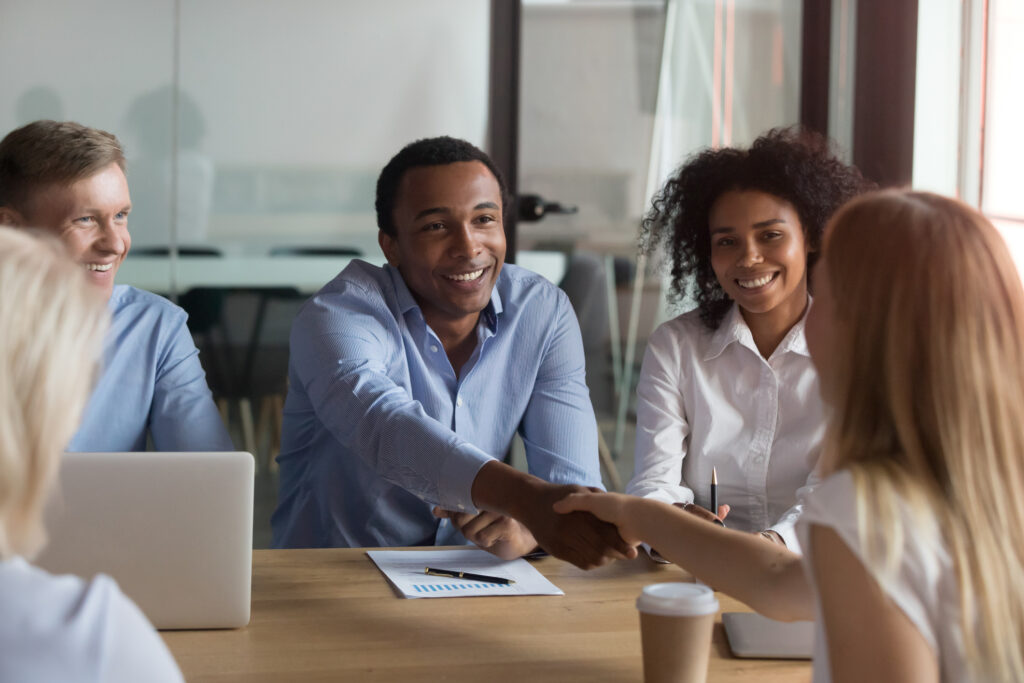 Successful diverse teammates sitting at boardroom, company head shaking hands with millennial specialist woman businesspeople ready to start conference discuss financial report planning future goals