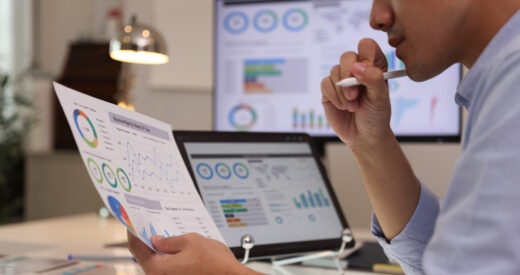 Focused businessman analyzing 2025 employee engagement trends reports on a desktop monitor and reviewing paper graphs, financial stats, and startup project infographics.