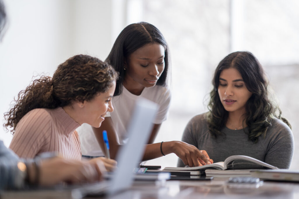 A young female University student of African decent, stands behind a peer and leans in as she tries to help her classmate with her studies. They are both dressed casually and are among a group sitting at the table all studying individually for class.