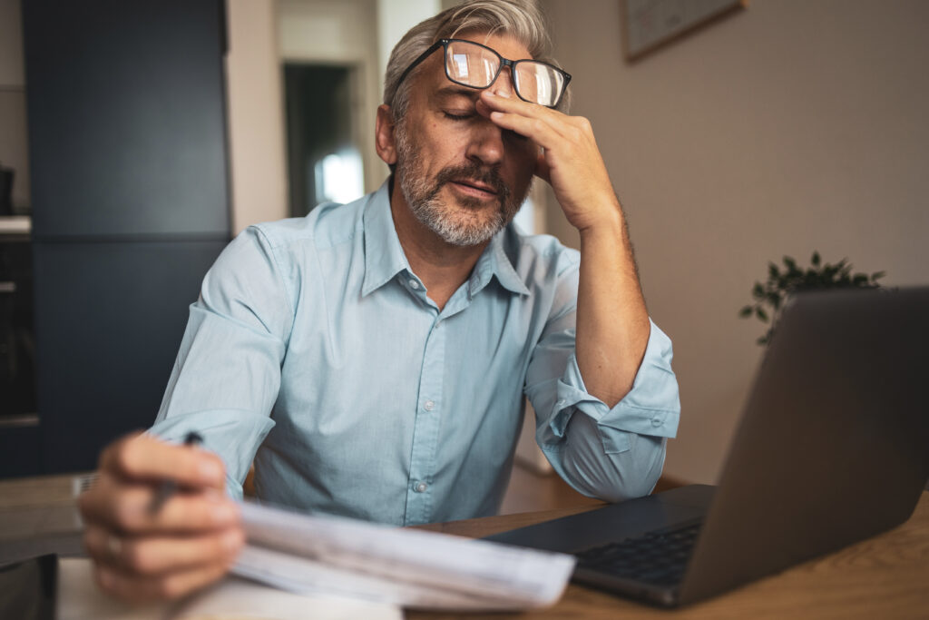 A mature man sits at home, reviewing paper financial statements while using a laptop. He appears thoughtful and focused, indicative of a serious stress approach to managing personal finances.