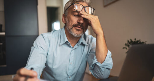 A mature man sits at home, reviewing paper financial statements while using a laptop. He appears thoughtful and focused, indicative of a serious stress approach to managing personal finances.